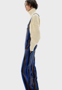 Salopette a righe verticali blu e marroni con tasche laterali, indossata sopra un maglione a collo alto color crema. Morbida al tatto, vestibilità rilassata e spalline regolabili.