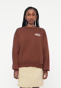 Sweatshirt castanho com um corte solto e mangas longas, apresentando o texto branco "New Balance Athletics" no peito. Combinado com uma saia bege.