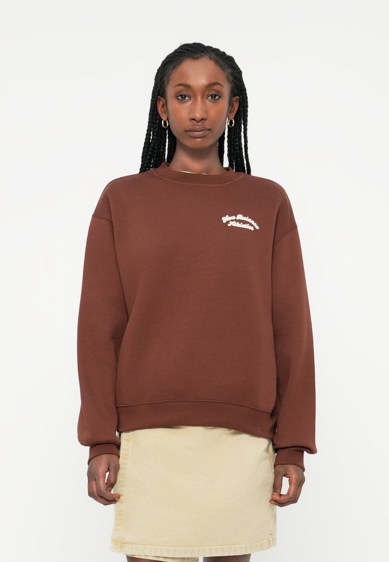 Brauner Sweatshirt mit lockerer Passform und langen Ärmeln, versehen mit weißem "New Balance Athletics"-Text auf der Brust. Kombiniert mit einem beigen Rock.