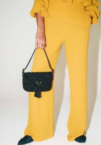 Borsa nera con perline e nappina, tenuta in mano. Indossata con pantaloni gialli a gamba larga e scarpe nere con punta affilata e design testurizzato.
