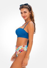 Blaue Bikini-Oberteil mit verstellbaren Trägern und strukturierter Stoff, kombiniert mit einem hochgeschnittenen Höschen mit einem bunten Blumenmuster auf einem cremefarbenen Hintergrund.