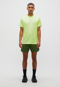 Νέος άνδρας που στέκεται με το πρόσωπο προς τα εμπρός, φορώντας ανοιχτό πράσινο αθλητικό μπλουζάκι, σκούρα πράσινα σορτς, μαύρες κάλτσες και μαύρα αθλητικά παπούτσια σε λευκό φόντο.