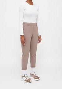 Pantalons ajustés de couleur beige clair avec une texture lisse, ourlets repliés et passants de ceinture, associés à un haut à manches longues blanc et des baskets claires.