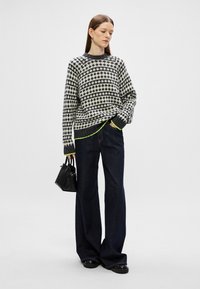 Pulover tricotat în alb și negru cu un model geometric, având un tiv inferior gri și accente de verde neon; purtat cu blugi negri, cu picioare largi, și o geantă neagră.