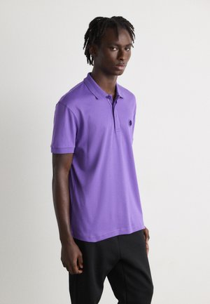 Purple polo shirt in soft fabric with a collar and short sleeves, featuring a small, dark logo on the left side. Black pants complete the outfit.