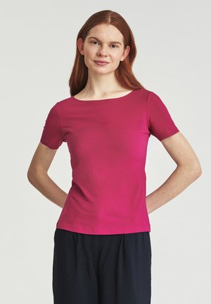 Giovane donna con lunghi capelli rossi che indossa una maglia rosa vivace a maniche corte e pantaloni scuri, in piedi con le mani dietro la schiena.