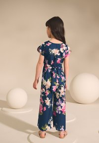 Cvetoč kombinezon iz temno modrega blaga z rožnatimi in belimi cvetovi, kratkimi nabranimi rokavi in pasom, ki se zaveže zadaj. Vidni so ravni sandali.