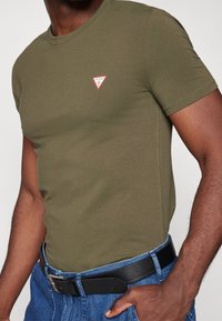 Mand iført en olivengrøn tætsiddende t-shirt med et lille rødt og hvidt logo på brystet, blå jeans og et sort læderbælte med sølvspænde.