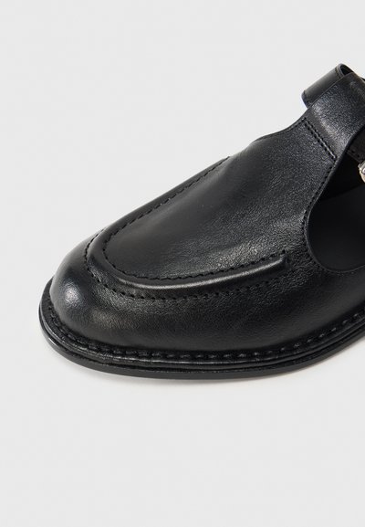 Μαύρο δερμάτινο παπούτσι με στρογγυλεμένη μύτη, ραμμένη λεπτομέρεια κατά μήκος των πλευρών και σχέδιο με κοπή. Διαθέτε ι κλείσιμο με λουράκι για ασφαλή εφαρμογή.