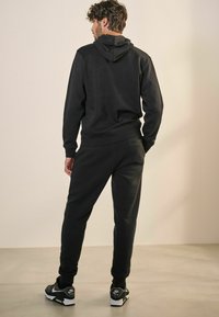 Svart hoodie med huva, ribbade cuffs och midja, ihop med matchande joggers. Modellen bär svarta sneakers med vita detaljer.