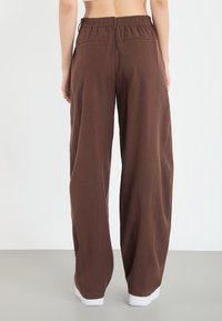 Pantalons amples marron en tissu doux, dotés d'une ceinture élastique et de deux poches arrière, reposant sur des baskets blanches.