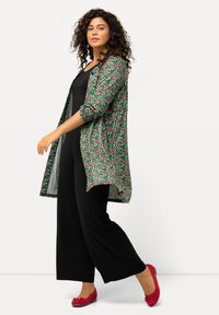 Cardigan lungo a fiori con sfondo verde e pois rossi e rosa, abbinato a una tuta nera e scarpe basse rosse. Tessuto dalla texture liscia.