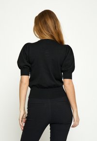 Vrouw met lang haar die van achteren wordt gezien, draagt een zwarte korte mouw trui met opgeblazen mouwen en nauwsluitende zwarte broek.