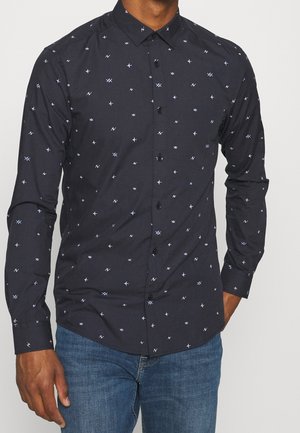 Uomo che indossa una camicia blu navy aderente con piccoli motivi astratti di croci e frecce azzurre, abbinata a jeans blu.