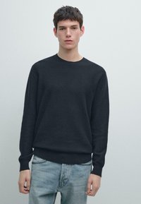 Maglione scuro in maglia con scollo rotondo e maniche lunghe, caratterizzato da un motivo a trama. Abbinato a jeans azzurri.