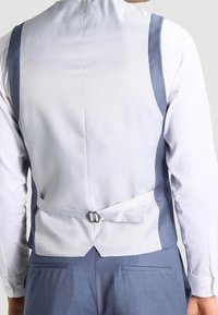 Chaleco gris claro con acentos azul oscuro, correa trasera ajustable, tela suave y diseño ajustado, combinado con una camisa blanca.