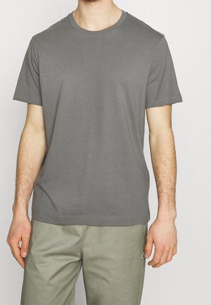 Mand iført en ensfarvet grå t-shirt med korte ærmer og lyse olivengrønne bukser, stående mod en neutral baggrund.