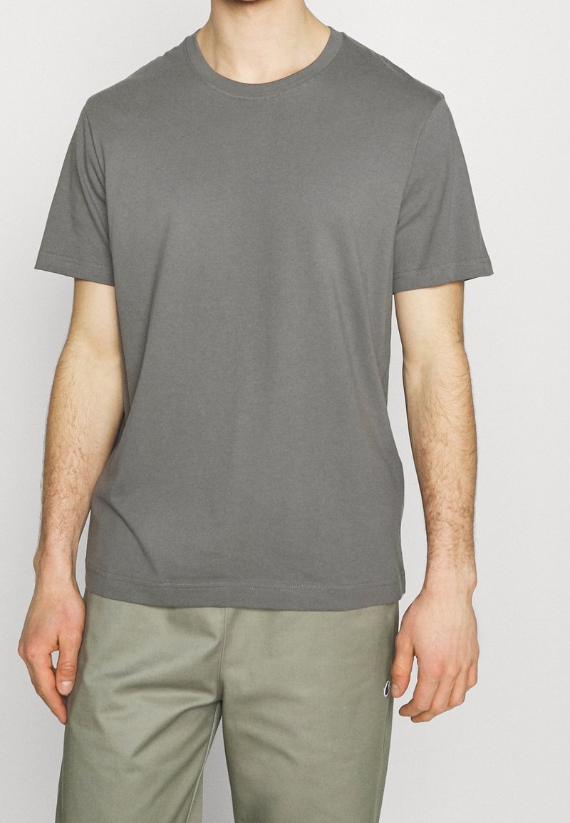 Homme portant un t-shirt gris uni à manches courtes et un pantalon vert olive clair, debout devant un fond neutre.