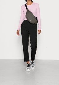 Sac bandoulière gris porté en travers du corps, avec une texture lisse et des fermetures éclair noires. Haut à manches longues côtelé rose clair et pantalon noir complètent la tenue.