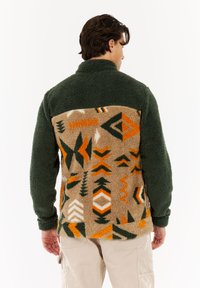 Mann trägt eine grüne Fleecejacke mit tanfarbenen, orangefarbenen und schwarzen geometrischen Mustern auf dem Rücken, kombiniert mit hellbeigen Cargo-Hosen, und steht mit dem Rücken zur Kamera.