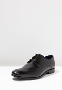 Chaussure habillée en cuir noir avec une finition lisse, bout arrondi, fermeture à lacets et un faible talon empilé. Détails de couture sur le côté visibles.