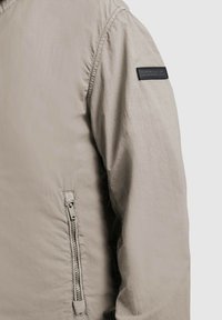 Svetlo béžový rukáv bundy s vreckom na zips pri páse a čiernou obdĺžnikovou náplasťou na hornej časti rukáva.