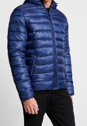 Mann trägt eine blaue gesteppte Steppjacke mit Frontreißverschluss und Reißverschlusstaschen, kombiniert mit schwarzer Hose vor einem einfarbigen Hintergrund.