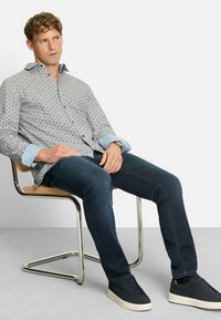 Mann mit lockigem Haar, der ein gemustertes Langarmhemd, dunkle Jeans und schwarze Sneakers trägt, sitzt auf einem modernen Metall- und Holzstuhl.