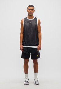 Νέος άνδρας που στέκεται βλέποντας μπροστά, φορώντας μαύρη αμάνικη αθλητική φανέλα, μαύρο αθλητικό σορτς, λευκές κάλτσες και ασπρόμαυρα αθλητικά παπούτσια σε απλό φόντο.