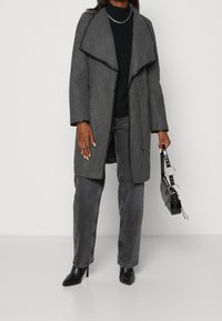 Vrouw in een lange grijze jas over een zwarte jurk en jeans, met een zwarte handtas met metalen ringhandvaten, die zwarte, puntige hakken draagt.