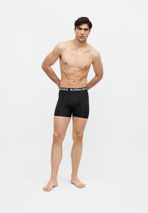 Uomo atletico in piedi a piedi nudi che indossa boxer neri Björn Borg con le mani dietro la schiena su uno sfondo chiaro e semplice.