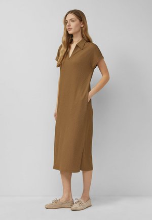 Kobieta stojąca bokiem, ubrana w luźną, brązową sukienkę do kolan z krótkim rękawem i beżowe buty casualowe, na jednolitym szarym tle.