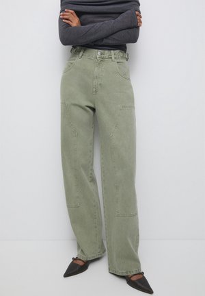 Person iført olivengrønne bukser med høj talje og vide ben, sorte spidse sko og en mørkegrå langærmet top med armene krydset.