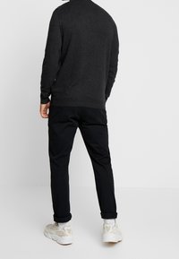 Dunkelgrauer Pullover mit langen Ärmeln, kombiniert mit schwarzen Hosen und hellen Sneakers. Die Rückansicht zeigt einen taillierten Schnitt und umgeschlagene Bündchen.