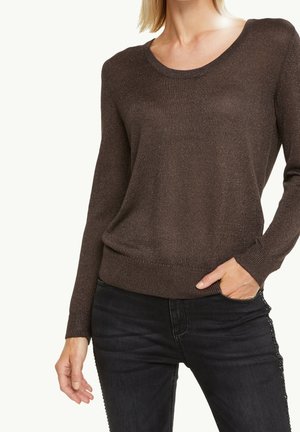 Kvinde iført en mørkebrun langærmet striktrøje og sorte jeans med den ene hånd i lommen, mod en ensartet lys baggrund.