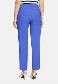 Pantalon bleu à jambes droites en tissu lisse, avec deux poches arrière. Porté avec des talons blancs montrant un accent noir sur la semelle.