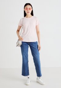 T-shirt de algodão rosa claro com mangas curtas, apresentando um logotipo discreto. Combinado com jeans de perna larga azuis e ténis brancos.