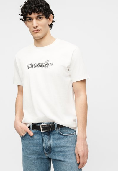 Νεαρός άνδρας με σγουρά μαλλιά, φοράει λευκό T-shirt με το λογότυπο Off-White, μπλε τζιν και μαύρη ζώνη, στέκεται με το ένα χέρι στην τσέπη.