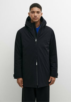 Young man wearing a black hooded zip-up coat over a blue shirt, standing against a plain light gray background.