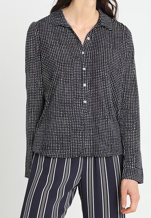 Femme portant une chemise noire à manches longues avec des pois blancs et un pantalon bleu marine à rayures verticales blanches et grises.