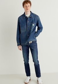 Giacca di jeans blu scuro e jeans con una t-shirt bianca sotto. Accentuata da una tasca frontale e chiusura con bottoni. Sneakers bianche.