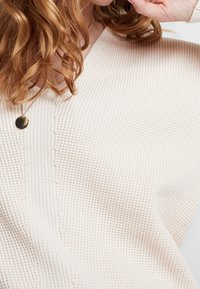Suéter de punto beige claro con textura de gofre, diseño de cuello en V y detalles de costura visibles. Lleva puesto un delicado collar dorado.