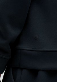 Sudadera negra con textura suave, corte relajado y un pequeño logo en relieve en la parte inferior delantera. Detalles de costura visibles.