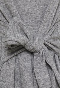 Měkká šedá pletená tkanina svázaná do velké, upravené uzly uprostřed, která ukazuje texturované švy a záhyby.