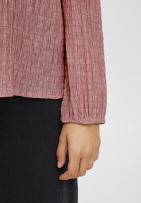 Rosa strukturierte Langarmbluse mit gerafftem Bündchen. Das Material wirkt leicht und zerknittert. Dunkelgraue Jeans sind teilweise sichtbar.