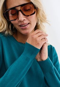 Kobieta w dużych brązowych okularach przeciwsłonecznych, srebrnych kolczykach w kształcie kół oraz turkusowym swetrze z długim rękawem, delikatnie składająca ręce.
