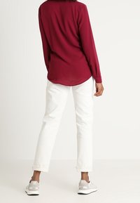 Bordeauxkleurige blouse met lange mouwen en een ronde zoom, gecombineerd met witte rechte jeans en grijze sportschoenen met lichte accenten.