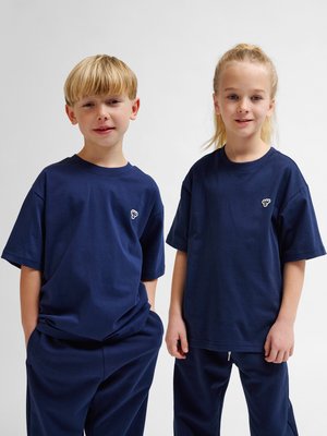 To børn står side om side iført matchende mørkeblå T-shirts og bukser mod en ensfarvet hvid baggrund.