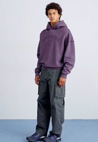 Junger Mann, der einen lila Kapuzenpulli, dunkelgraue Cargo-Hosen und schwarze Turnschuhe trägt, steht vor einem schlichten hellen Hintergrund.