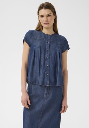 Žena v modrom krátkom rukáve džínsy top s gombíkmi a zodpovedajúcu džínsy sukňu, stojaca proti jednému svetlému pozadiu.